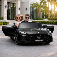 Two children sitting in a black 24 v Mercedes-Benz AMG toy car on a driveway.