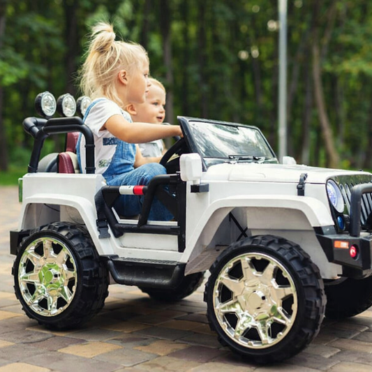 Two small blond children sitting in an electric ride-on SUV. They are driving over a brick path lined with trees.
