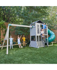 Kidkraft Wrangler Way Swing Set, Wooden Playhouse & Slides Children playing on a playground set with a swing, slide, and playhouse in a backyard.