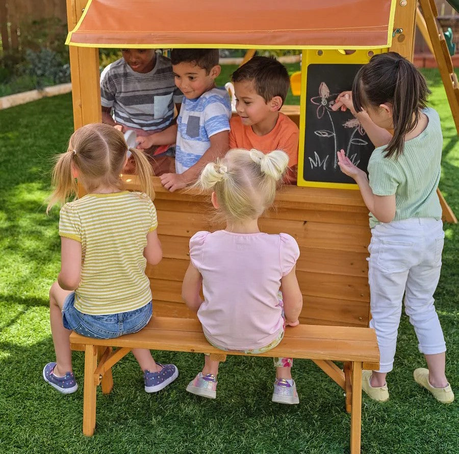 Kidkraft Overland Heights Children playing around a wooden playhouse with a chalkboard on a grassy area.