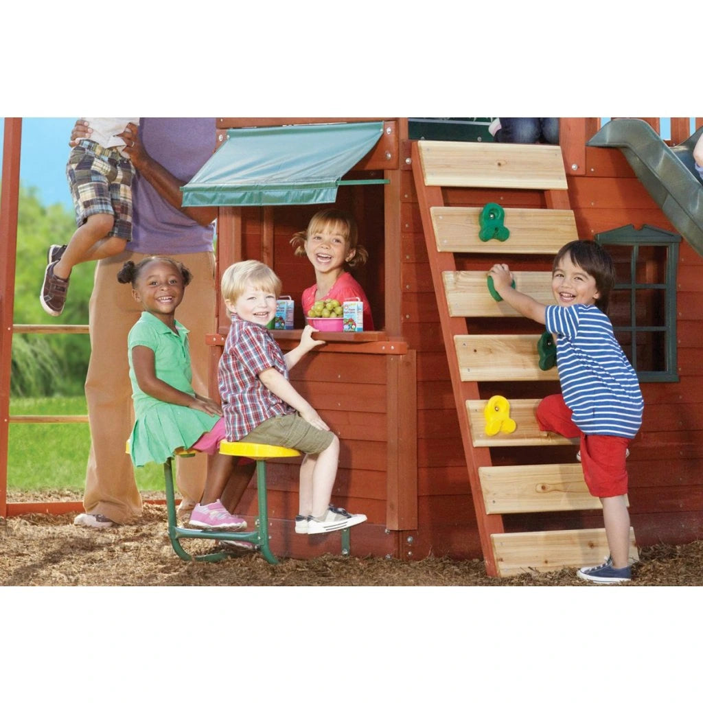 KidKraft Ridgeview Deluxe Clubhouse Children playing on a wooden playhouse with a slide and climbing structure.