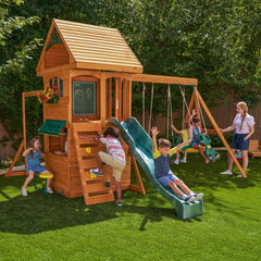 KidKraft Ridgeview Deluxe Clubhouse Children playing on a wooden playset with a slide and swings in their backyard.