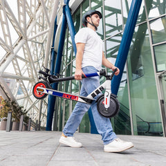 Person carrying a Folable E scooter in an urban setting with modern architecture.