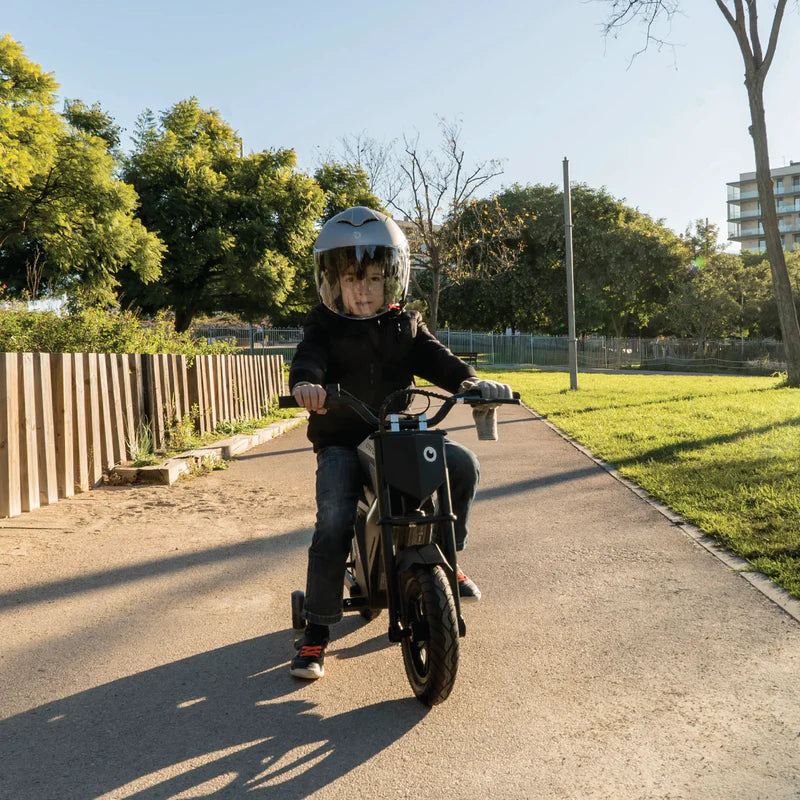 Child riding a small motorcycle on a path with trees and buildings