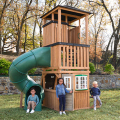 KidKraft Panorama Tower Outdoor Playhouse Children playing on a wooden playhouse with a green slide in a backyard setting.