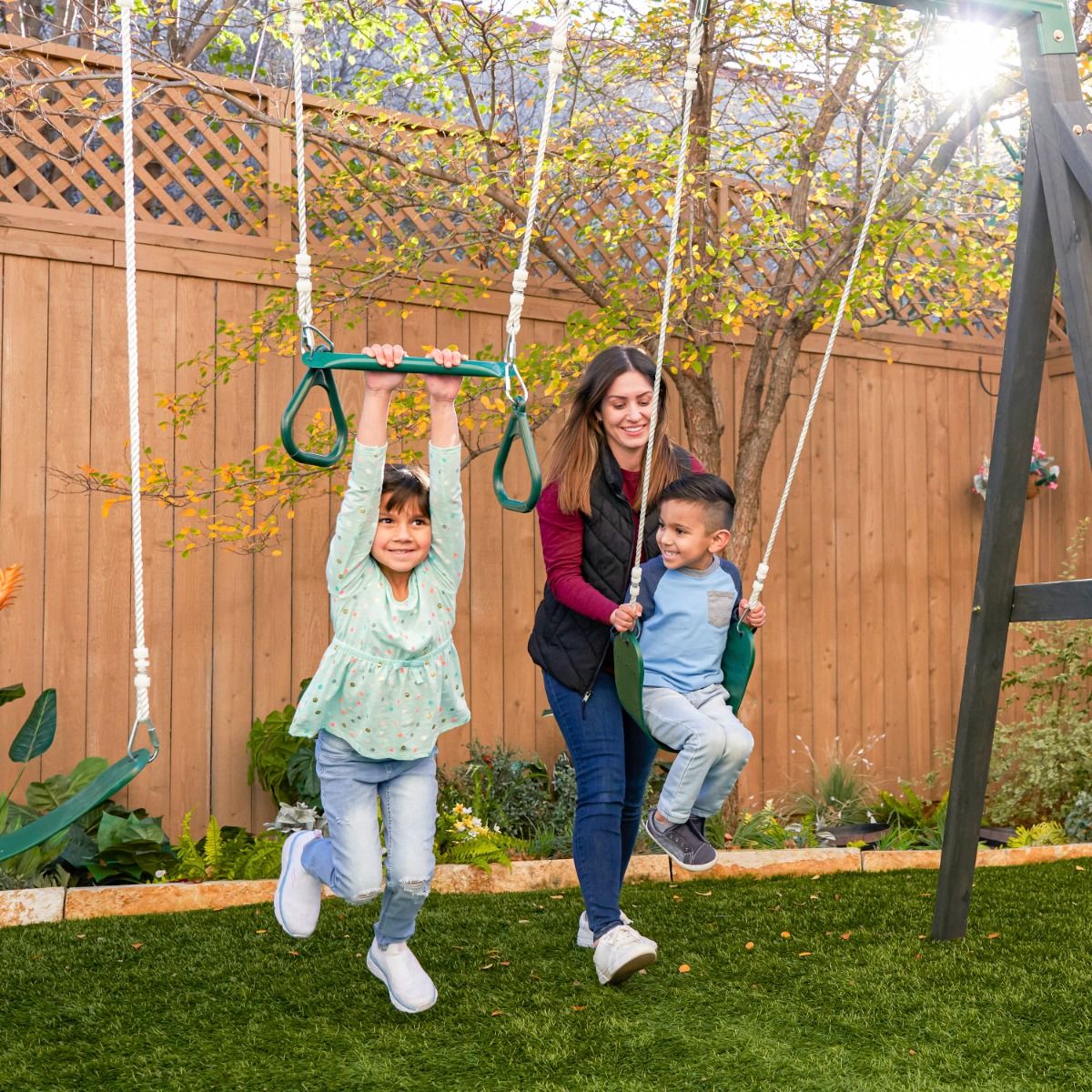 Kidkraft Wrangler Way Swing Set, Wooden Playhouse Woman and two children on a swing set in a backyard