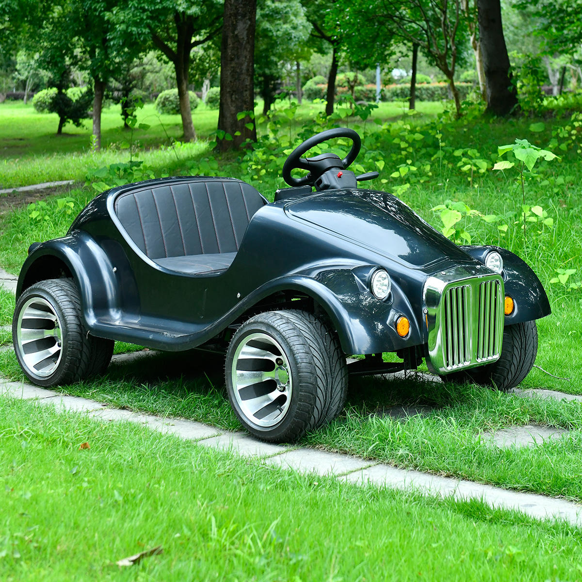 A black vintage-style off road gasoline convertible ride-on car with chrome-like silver wheels, parked on a grassy path in a park.