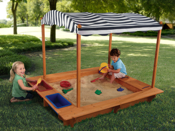 Children playing in a sandbox with a striped canopy on a grassy playhouse.